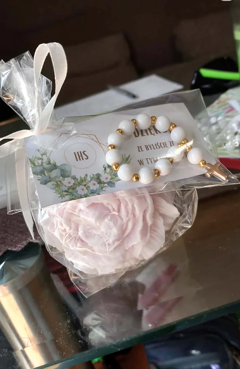 Gift package containing a white and gold beaded religious bracelet with IHS charm, decorative card, and pink flower, wrapped in cellophane